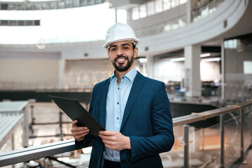 Construction engineer smiles while holding a clipboard at a modern construction site. The location...