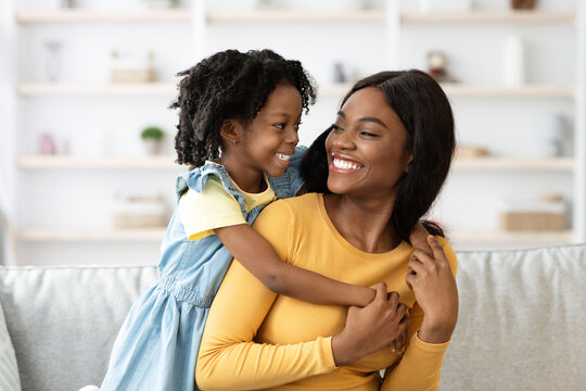 A young girl smiles brightly while hugging her mother from behind. Both display happiness and warmth in a cozy home setting. They enjoy quality time together.