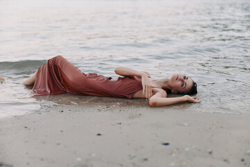 Woman lying on sandy shore in a flowing red dress, relaxed pose as gentle waves lap at her side, creating a calm, ethereal beach scene.