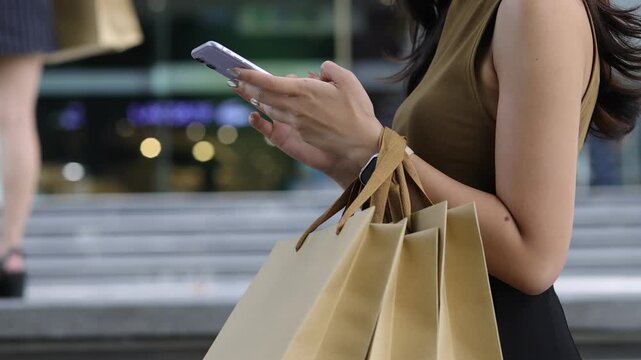 Female tourist walking city streets, carrying multiple shopping bags while browsing smartphone, experiencing digital retail therapy with evident enjoyment and urban lifestyle satisfaction.