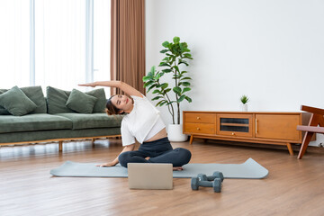 Young Asian woman stretching yoga workout on exercise mat while online training class with computer laptop in living room