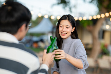 Asian woman clinking alcohol drink bottle with male friend sitting at table at campground restaurant