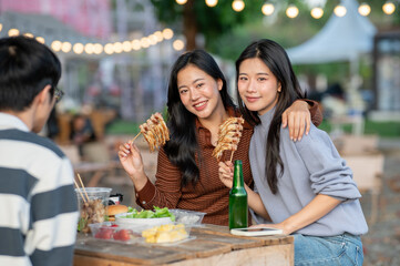 Asian woman eating meat skewer and hugging friend while sitting at table in a campground restaurant.