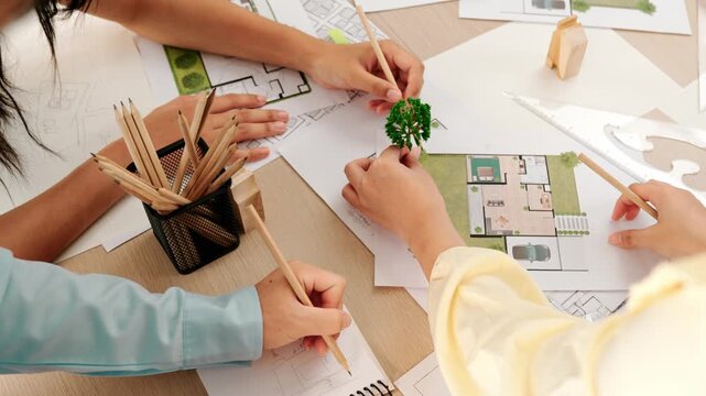 Architect and engineer use tree model discuss about building blueprint on table with architectural document, measurement tools and wooden block scatter around at modern office. Closeup. Delineation.