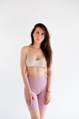 Fototapeta premium Sports woman standing in activewear with abs muscles, looking at camera isolated on white background. Caucasian 30s fitness girl wears beige top, pink leggings. Happy fit smiling brunette, blue eyes.