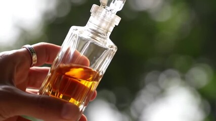 Hand holding a vintage perfume bottle with amber liquid against a blurred green background. - Powered by Adobe