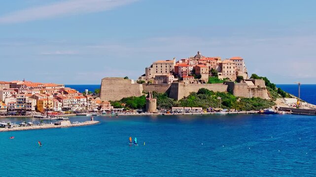 Aerial drone shot over the coastal town of Calvi in Corsica. View of the citadel fortress overlooking the city. Summer holidays destination. Bright blue sky, vibrant turquoise sea.