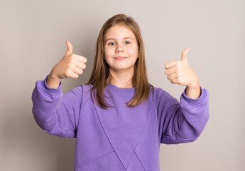 Smiling preteen girl in purple outfit showing thumbs-up gesture on grey background. Perfect for approval, success, positivity, encouragement, or achievement concepts.