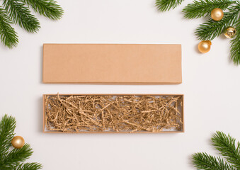 Open long kraft gift box with paper filler on white background decorated with fir branches and golden mini balls. Perfect for Christmas packaging, eco gift, or holiday presentation concepts.
