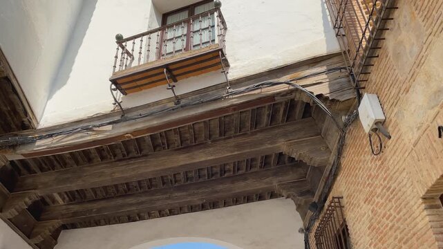 Slow-motion upward sequence of an exterior corridor connecting two buildings in La Puebla de Montalb&aacute;n. Wooden beams, whitewashed walls, brick, and stone textures enhance the traditional charm.