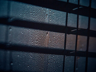 Close view of window with dark blinds, condensation and raindrops in focus, soft morning light reflecting from the right, cozy atmospheric interior, warm matte tones, intimate and moody composition.