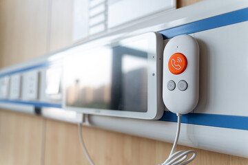 A medical call device with an orange button and a screen is mounted on a wall in a hospital room.