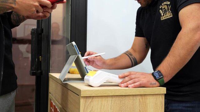Handheld shot of customer placing phone over dataphone for contactless payment while barber confirms transaction on tablet with stylus
