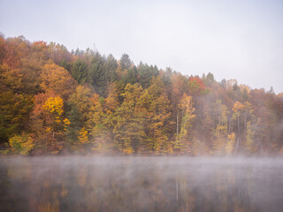 Colorful autumn forest reflected on calm lake surface with morning mist. Peaceful nature scene from Ružín reservoir, Slovakia.