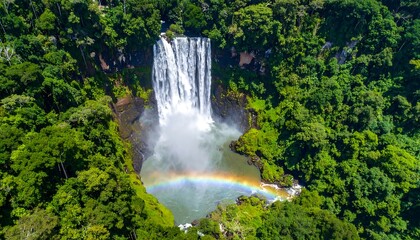 Obraz premium Cascading waterfall plunges through lush green forest foliage, with rainbow forming at the base creating mist