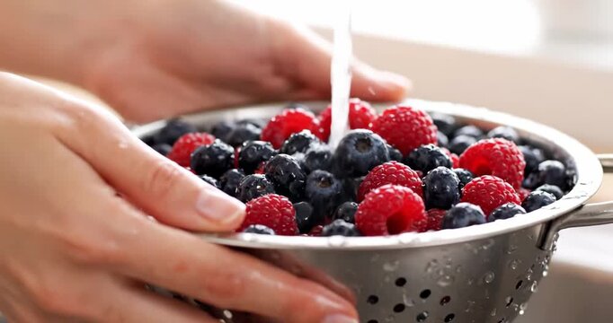 Washing berries in a colander, medium close-up view of hands gently rinsing a colander full of fresh blueberries and raspberries under gently running water.