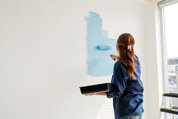 Person painting a white interior wall light blue with a roller near a window, holding a paint tray during a home makeover