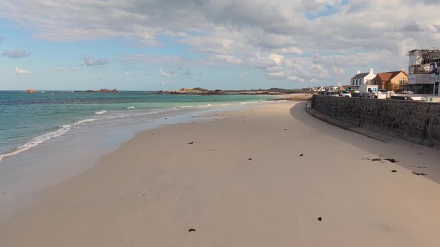 Low close in drone footage of shoreline at Cobo Bay Guernsey Channel Islands with sandy beach clear turquoise water lapping waves and showing beachfront properties on a sunny day with interesting sky