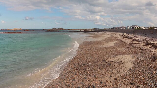 Low forward drone footage of shoreline at Cobo Bay Guernsey Channel Islands with pebbles on sandy beach clear water and lapping waves on a sunny day with interesting sky