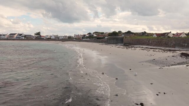 Low close in drone footage of shoreline at Cobo Bay Guernsey Channel Islands with deserted sandy beach clear water lapping waves and sea birds with interesting sky