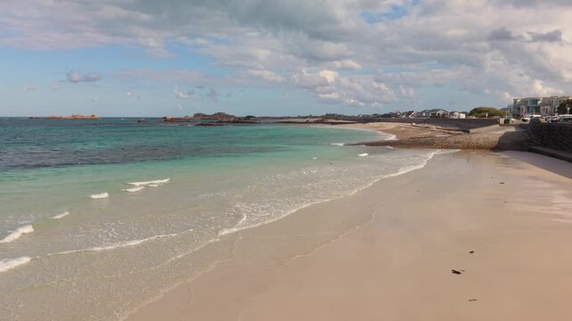 Low drone footage of shoreline at the stunning Cobo Bay Guernsey Channel Islands with deserted sandy beach clear turquoise water and lapping waves on a sunny day with interesting sky