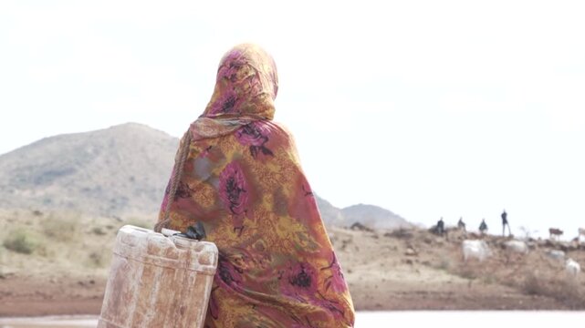 African woman carrying a jerrycan walking towards a dirty water pond.
