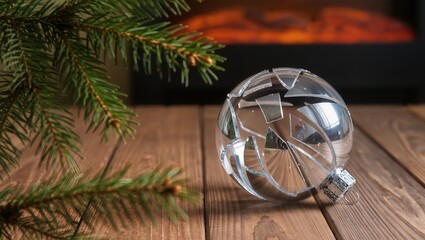 Top view of a cracked silver Christmas ornament on a wooden floor with warm light, blurred pine branches, and a glowing fireplace in the background
