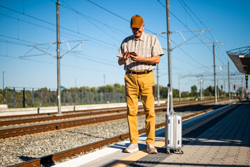 Happy senior man is standing at railway station and waiting for arrival of the train. He is using smartphone. Senior people traveling.