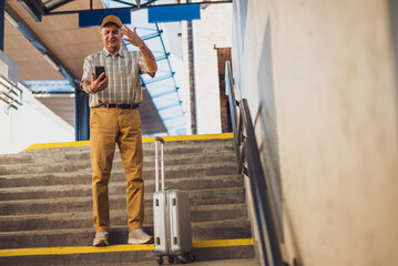 Senior man is standing at railway station and messaging on smartphone. He is distraught. Senior people traveling.