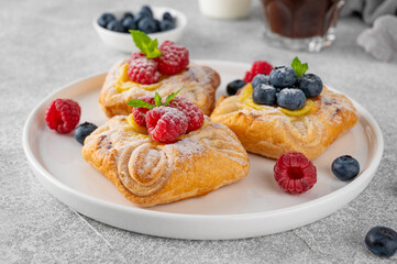 Danish puff pastry cakes with custard cream and fresh berries, sprinkled with powdered sugar on a white plate on a concrete background.