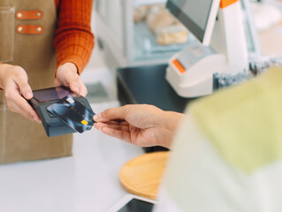 Close up of Customer hand using credit card tapping on Card Reader, terminal for making payment in coffee shop. Cashless and Contactless payment buying drink, making payment transaction to cashier.