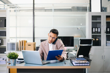 Fototapeta premium Asian businessman talking on smartphone while working on laptop at modern office desk, symbolizing business communication and corporate success.