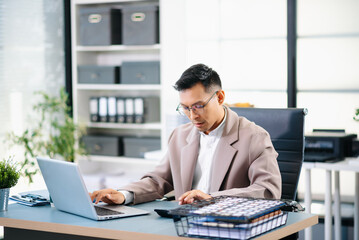 Obraz premium Confident Asian businessman standing at desk, reviewing report and working on laptop in modern office