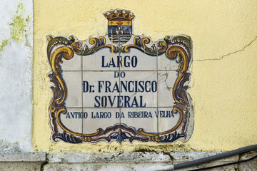 Ceramic tile sign bordered with ornate blue and gold designs, featuring a crown and coat of arms at the top, with text indicating the name and historical reference of the location in Faro, Portugal