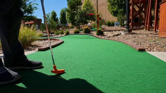 Playing mini golf with a yellow ball and orange putter on green astroturn on a sunny day - shot from behind golfer