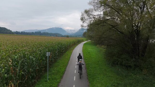 Aerial: couple riding e-bikes in the countryside along Drava River with moody sky during the day, tracking drone shot