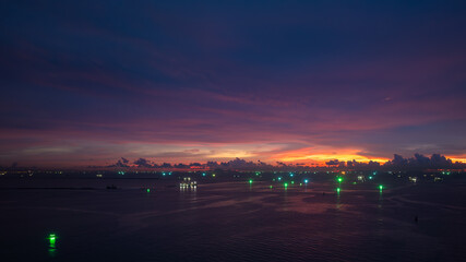 twilight time over the sea, The sunset sky is filled with deep purple, orange, and pink hues. Bright green lights from fishing and other boats twinkle on the water,