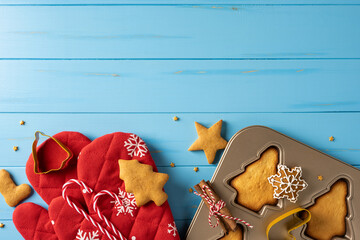 Festive holiday baking scene featuring red Christmas mitts, star and snowflake cookies, candy canes, and a baking tray on a blue wooden surface, ready for treats and celebrations