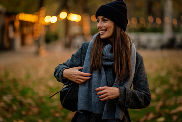 Confident and attractive woman wearing warm knitted scarf and beanie while standing in the park in...