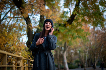 Stylish middle-aged woman wearing long coat and knitted beanie and using smartphone outdoors