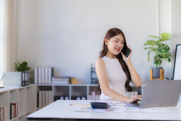 Young Asian woman sitting at her office desk, effectively multitasking by typing on a laptop and engaged in a phone conversation, portraying efficient business communication and productivity