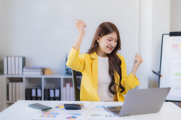 Asian businesswoman in modern office celebrates career milestone, arms raised over laptop showing growth charts and analytics, smiling confidently at financial success
