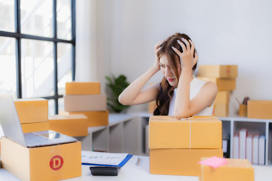 Businesswoman feeling stressed and frustrated, holding her head in her hands, sitting at a desk full of shipping boxes and a laptop, struggling with the demands of her online small business
