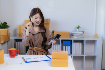 Young Asian woman working from home, calculating financial data and managing orders for her online e-commerce business, surrounded by shipping boxes and office supplies