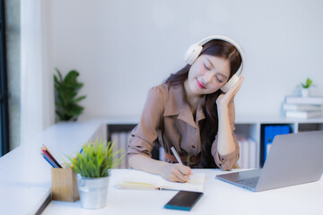 Young woman in headphones smiling as she studies and works at a laptop, writing notes at a modern office desk, relaxed, focused and productive in a calm workspace