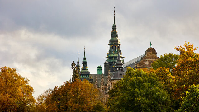 Historic Nordiska Museet building in Stockholm surrounded by colorful autumn trees under a dramatic cloudy sky. Scenic view of famous Swedish architecture during fall season.
