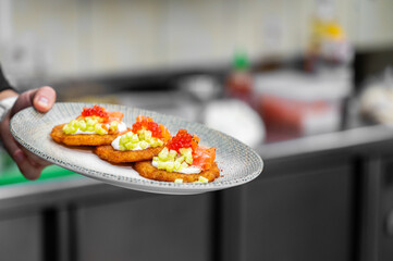 Gourmet potato pancakes topped with sour cream, smoked salmon, red caviar, and fresh herbs on a rustic plate. A chef's hand is visible holding the plate