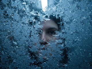 A person peers through a window frame covered in intricate blue frost and ice crystals, creating a cold, mysterious portrait.
