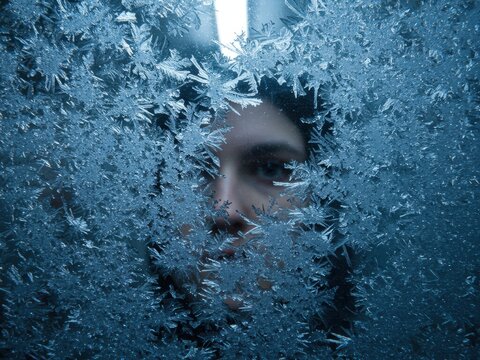 A person peers through a window frame covered in intricate blue frost and ice crystals, creating a cold, mysterious portrait.