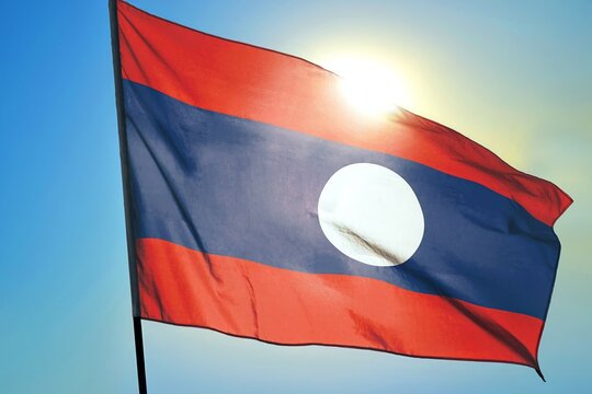 Laos national flag waving in sunlight close-up – Lao red blue red horizontal stripes banner with white circle patriotic symbol of Southeast Asia background blue sky bright sunny day